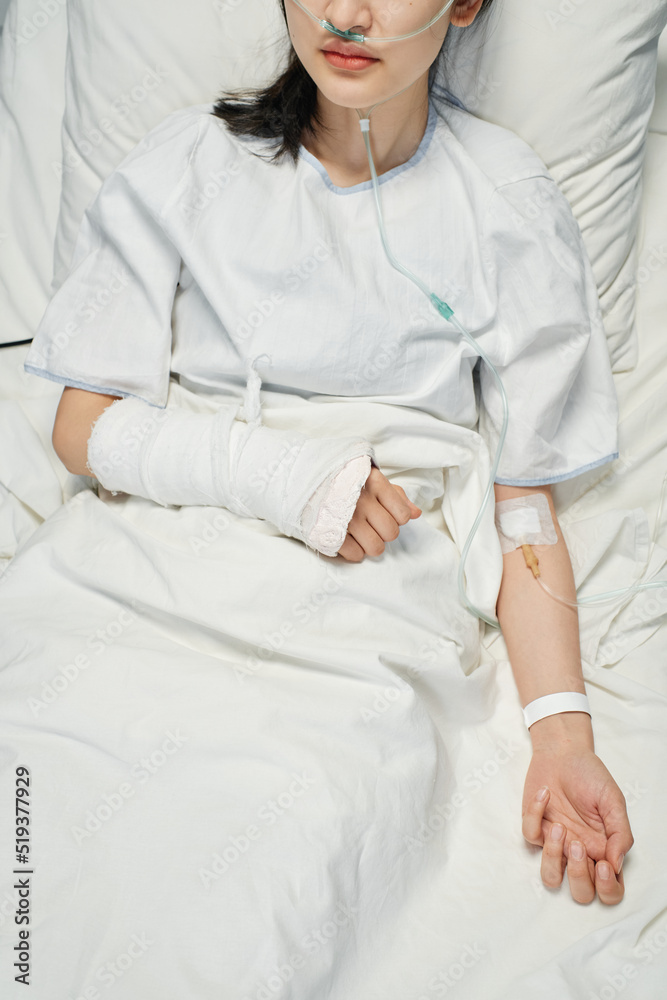 Vertical high angle shot of young Asian patient with broken arm in cast receiving medication or nutrient fluids through intravenous line