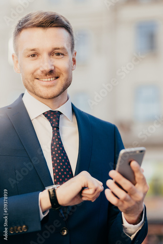 Smiling manager director with watch on arm, dressed elegantly, holds smart phone, reads positive news about his new project, poses outdoor, waits for colleague near office building. Job concept photo