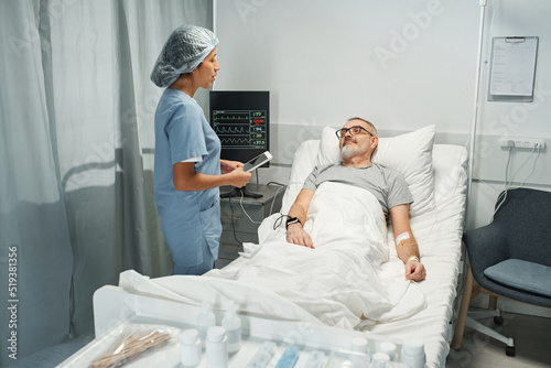 Young adult medical worker holding digital tablet talking to mature man checking his health state in emergency room