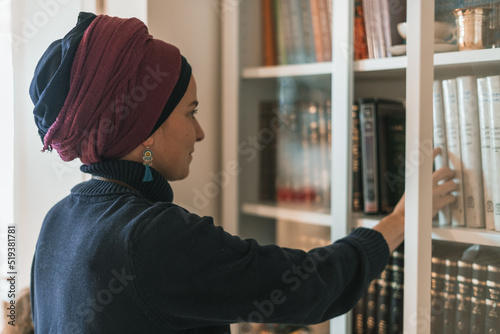 Religious jewish young woman with head covered stands near bookcase with religious books (9)