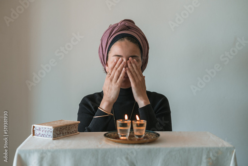 Jewish woman prays over lit Shabbat candles, covering her face with her hands. Nearby lies a religious prayer book. Jewish religious traditions (71)