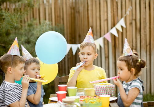 Cute funny nine year old boy celebrating his birthday with family or friends with homemade baked cake in a backyard. Birthday party. Kids wearing party hats and blowing whistles.