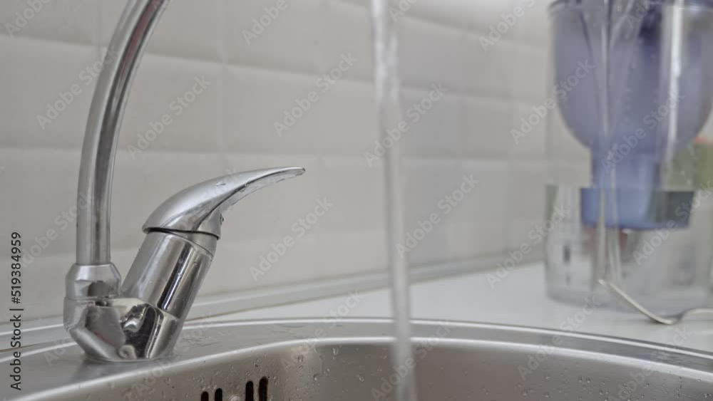A man in a black jacket opened a tap with water in the kitchen against the background of a white wall, The man turned on the water in the sink. Close-up.