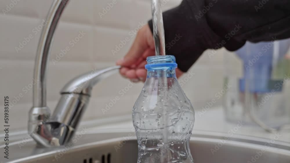 Close-up of a man in a black jacket pouring cold tap water into a plastic bottle in a home sink in the kitchen against a white wall. Close-up of a man in a black jacket pouring cold tap water into a plastic bottle in a home sink in the kitchen against a white wall.