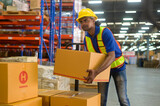 Young mixed race male worker wearing helmet lifting cardboard box in warehouse, machinery and Logistics concept.
