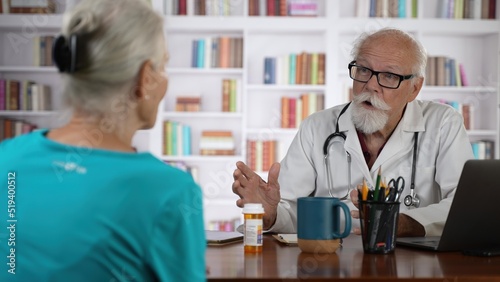 Mature senior male doctor talking to woman patient in modern clinic with library background.