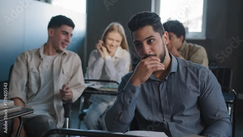 Sad frustrated arab student sitting in class at desk alone suffering from mistreatment by classmates racial discrimination feels victim of bullying violent students bully ridicule gossip behind back