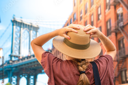 Fashionable tourist woman visiting New York City wearing hat and stylish street style outfit. Young girl amazed by the beautiful city. Manhattan bridge view on summertime.