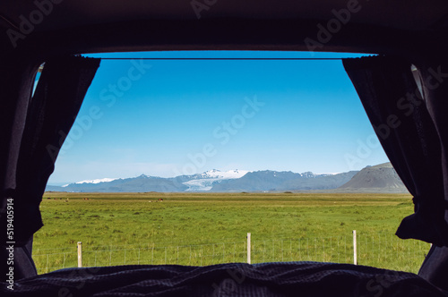 Looking out the camper minivan back from built in bed. Traveling Iceland with van. View over mountains, pasture and glacier, blue sky, sunny day.