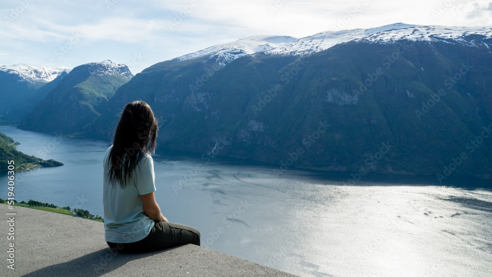 Unrecognizable woman sitting on the edge of a lookout point with the fjord in the background in Norway.