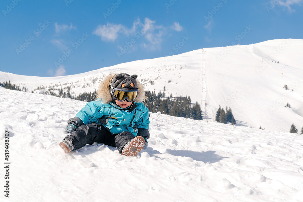 Little cute baby kid child boy in winter overalls,ski mask,glasses goggles sitting on snow at winter holiday, in ski resort in Carpathian high mountains outdoor nature landscape, Ukraine, Europe Little cute baby kid child boy in winter overalls,ski mask,glasses goggles sitting on snow at winter holiday, in ski resort in Carpathian high mountains outdoor nature landscape, Ukraine, Europe