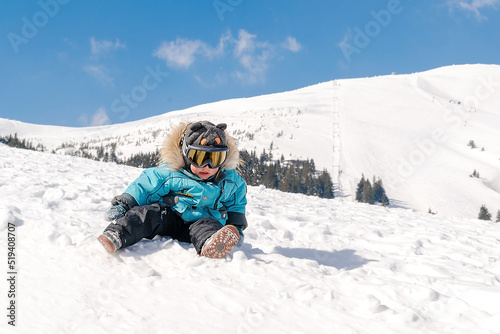 Little cute baby kid child boy in winter overalls,ski mask,glasses goggles sitting on snow at winter holiday, in ski resort in Carpathian high mountains outdoor nature landscape, Ukraine, Europe