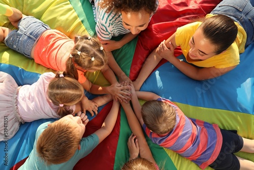 Group of children with teachers holding hands together on rainbow playground parachute, top view. Summer camp activity photo