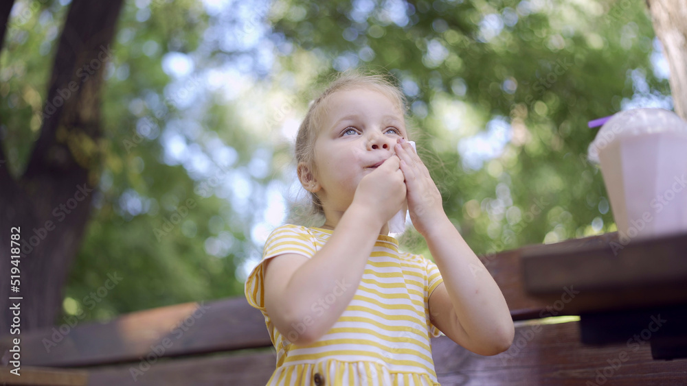 Naklejka premium Little girl wipes her face with a paper napkin after eating, there is a milkshake on the table. Close-up of cute child girl sitting on park bench and wiping her face with a paper napkin.