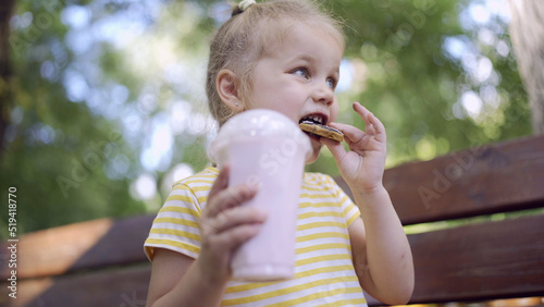 ittle girl eats a colorful gingerbread and holds a milkshake in her hand. Close-up of cute child girl sitting on park bench and eating cookies with a milkshake.