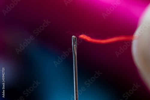 A portrait of a metal sewing needle with someone trying to put a red thread through the eye of the piece of sewing equipment. © Joeri