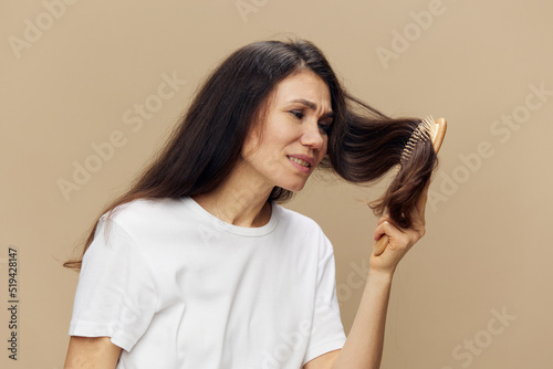 portrait of a beautiful woman in a white T-shirt on a beige background combing her long hair with a wooden massage comb and twisting her face in pain