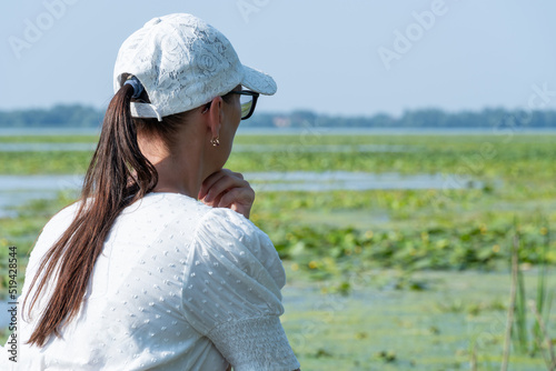 Darkhaired woman looks thoughtfully at the overgrown river. She is wearing white blouse and cap. Slowly flowing river is overgrown with yellow water lilies. Ecological problem.