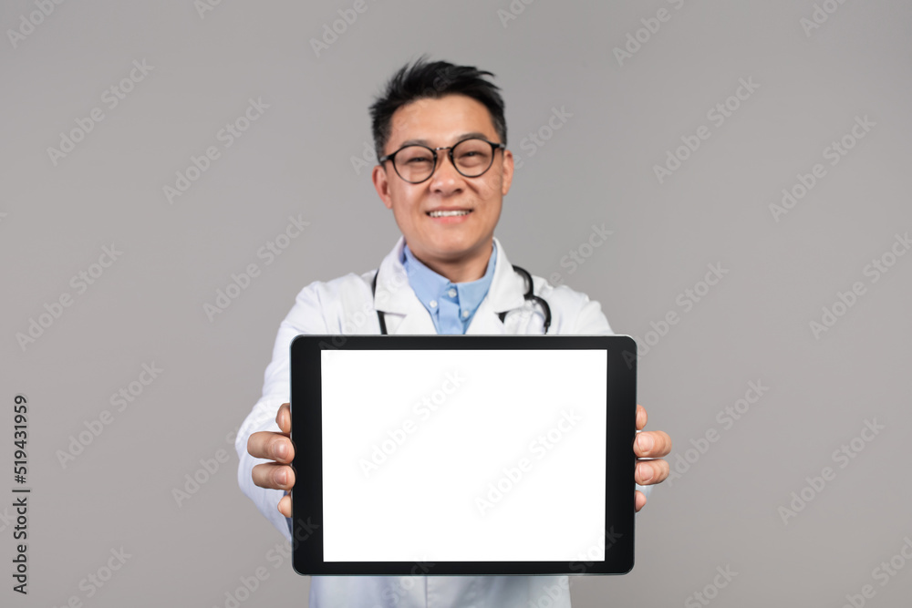 Happy confident adult japanese doctor in white coat and glasses shows tablet with blank screen Happy confident adult japanese doctor in white coat and glasses shows tablet with blank screen