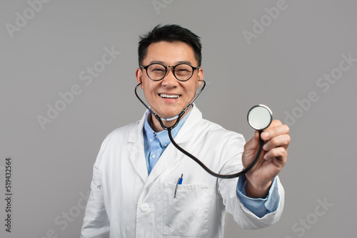 Cheerful confident mature japanese doctor in white coat and glasses listens camera with stethoscope