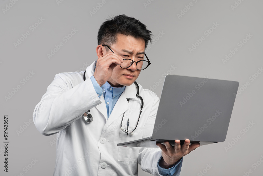 Confused concentrated adult asian man doctor in white coat takes off glasses, looks at laptop Confused concentrated adult asian man doctor in white coat takes off glasses, looks at laptop