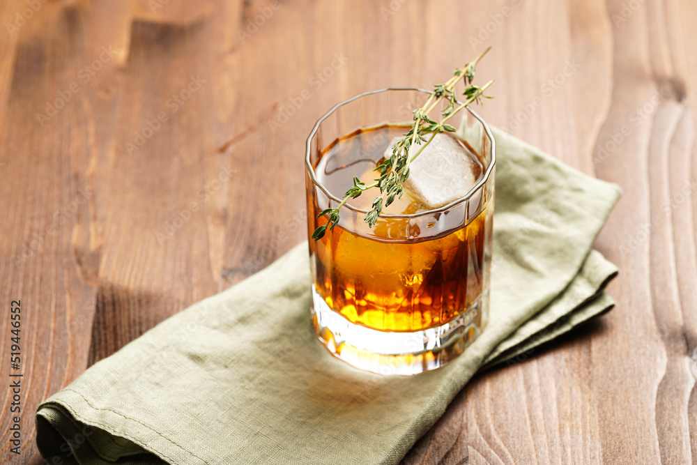 a tumbler glass with brown alcoholic drink, thyme and ice cubes on linen napkin - whisky, rum or cognac - on wooden table, rural scene a tumbler glass with brown alcoholic drink, thyme and ice cubes on linen napkin - whisky, rum or cognac - on wooden table, rural scene