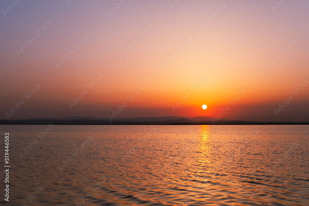 Colorful orange, yellow and purple sunset over water of lake with sun setting in the water. Natural park at sunset with distant silhouettes of mountains. Sunset landscape view from a boat. Traveling 