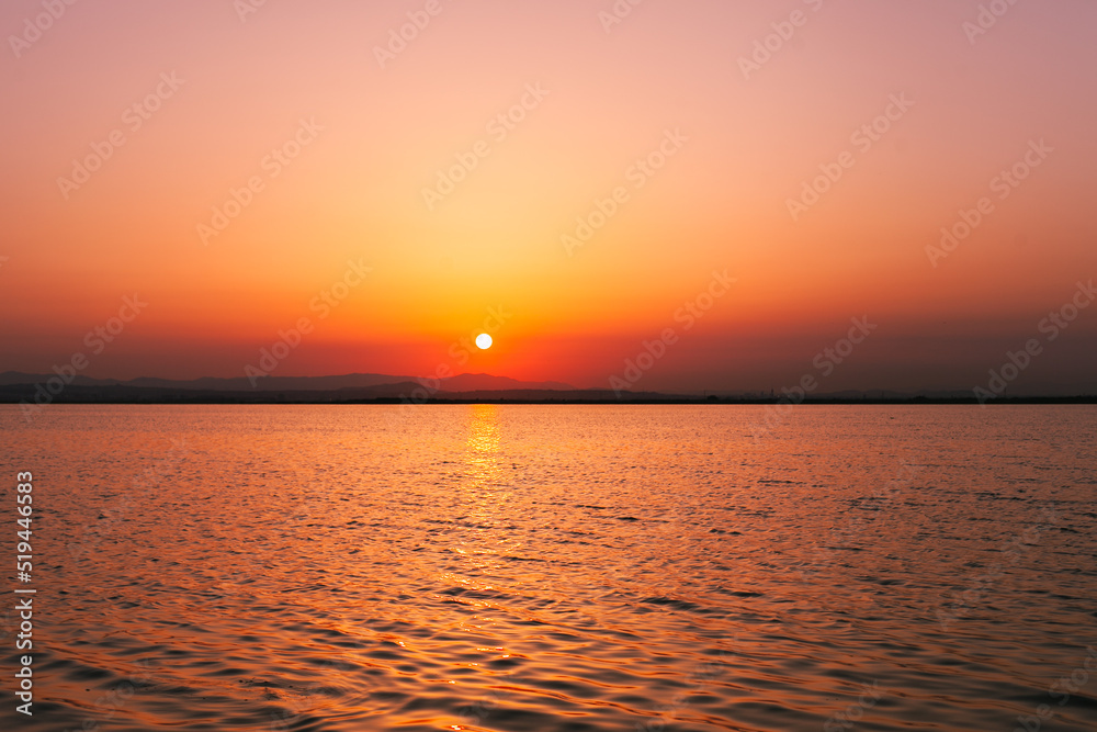 Colorful orange, yellow and red sunset over the water of lake with sun setting in the water. Natural park at sunset with distant silhouettes of mountains. Sunset landscape view from a boat. Traveling 