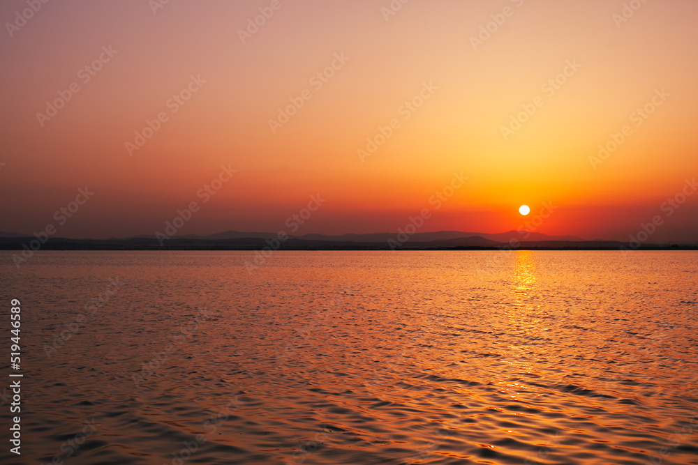 Colorful orange, yellow and red sunset over the water of lake with sun setting in the water. Natural park at sunset with distant silhouettes of mountains. Sunset landscape view from a boat. Traveling 