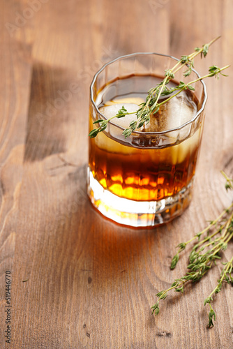 a tumbler glass with brown alcoholic drink, thyme and ice cubes - whisky, rum or cognac - on wooden table, rural scene