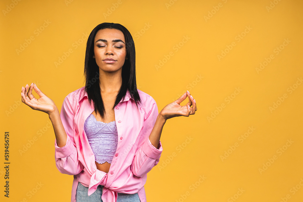 Meditation concept. Beautiful young african american woman stands in meditative pose, enjoys peaceful atmosphere, holds hands in praying gesture, isolated over yellow background. Meditation concept. Beautiful young african american woman stands in meditative pose, enjoys peaceful atmosphere, holds hands in praying gesture, isolated over yellow background.