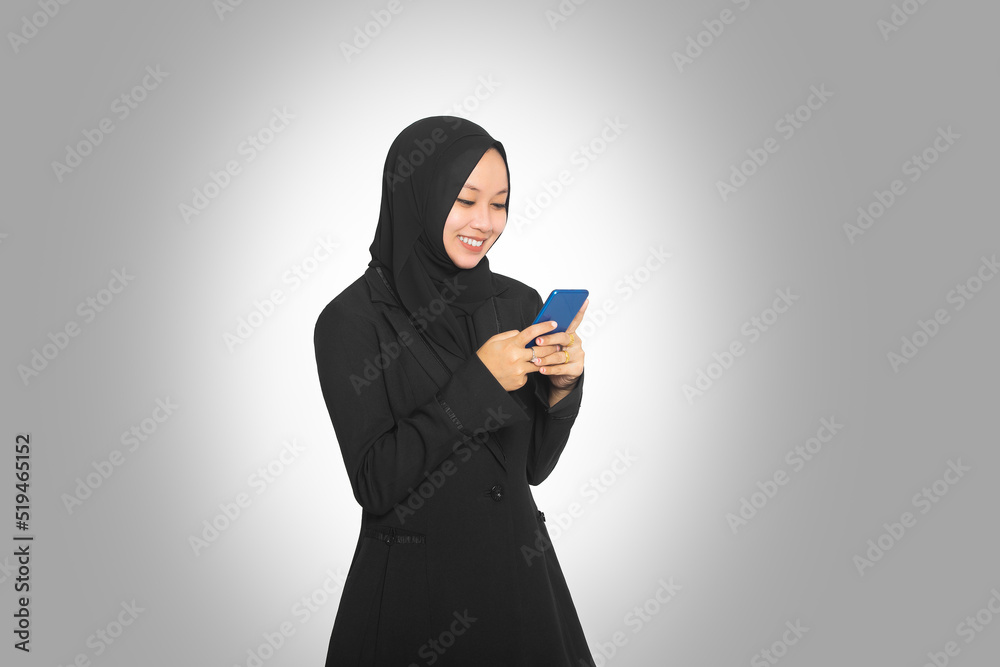 Beautiful young Muslim business woman wearing hijab holding smartphone in hand smiling standing isolated on gray background Beautiful young Muslim business woman wearing hijab holding smartphone in hand smiling standing isolated on gray background