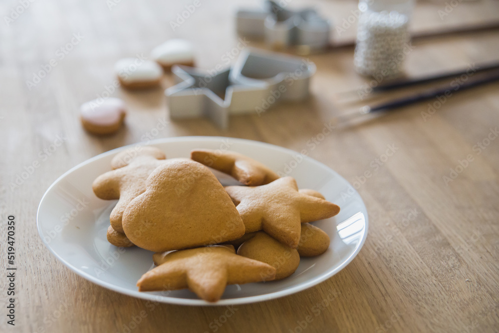 A white plate of traditional ginger biscuits stands on a wooden table. Brushes for drawing on gingerbread, forms for gingerbread in the form of hearts and stars. A white plate of traditional ginger biscuits stands on a wooden table. Brushes for drawing on gingerbread, forms for gingerbread in the form of hearts and stars.