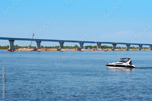 A small speedboat floats along the river against the backdrop of a bridge under construction.