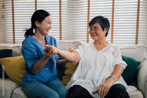 Delightful female caregiver in blue uniform smiling and stretching arm of elderly Asian woman while sitting on sofa against window in living room at home