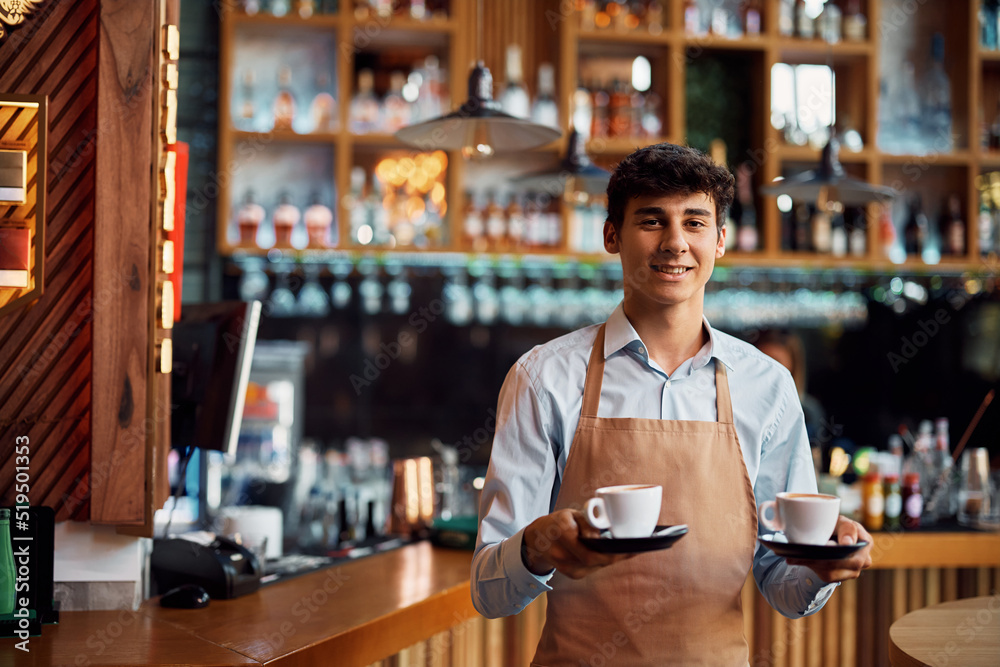 Young waiter serving coffee in cafe and looking at camera.