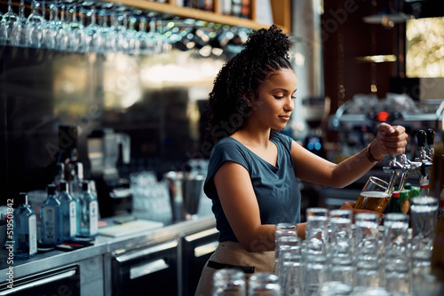 Young black waitress pours beer draft beer while working at bar counter. photo