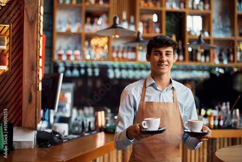 Obraz na plátně Young waiter serving coffee in cafe and looking at camera.