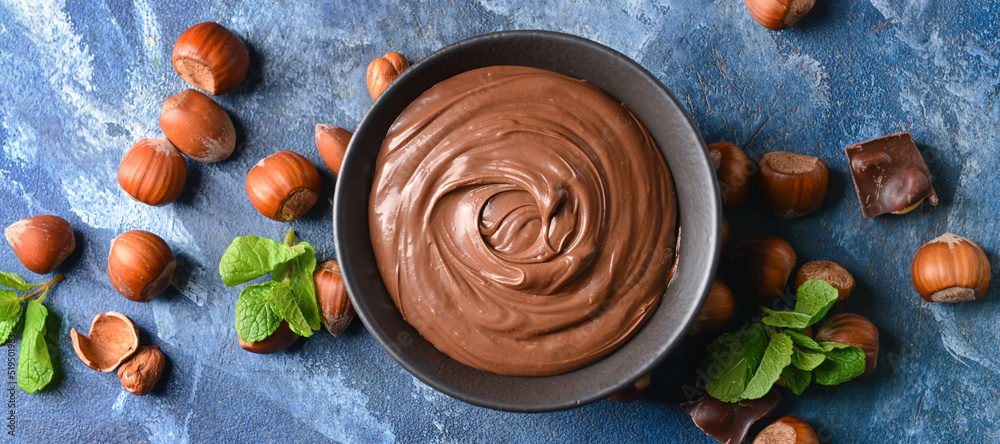 Bowl with tasty chocolate paste and hazelnuts on blue background, top view