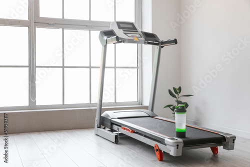 Modern treadmill with bottle of water and houseplant near window in room