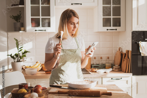 Pretty young woman preparing apple pie at home in cozy kitchen and takes pictures on phone