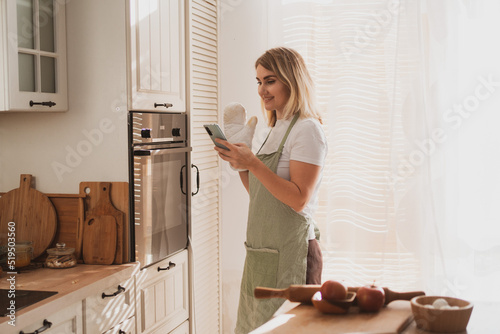 charming young woman in chef's apron puts apple pie in the oven and takes pictures on phone