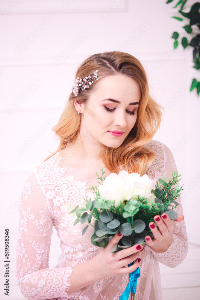 Young attractive bride with blond hair with wedding makeup and hairstyle in a white lace peignoir with bride's bouquet in a bright interior Young attractive bride with blond hair with wedding makeup and hairstyle in a white lace peignoir with bride's bouquet in a bright interior