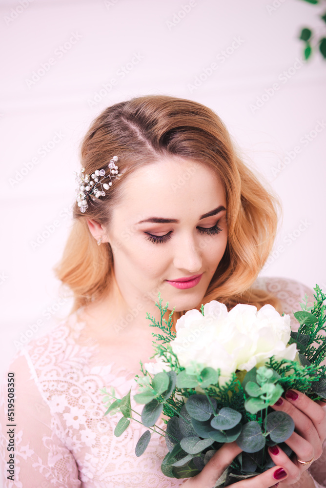 Young attractive bride with blond hair with wedding makeup and hairstyle in a white lace peignoir with bride's bouquet in a bright interior Young attractive bride with blond hair with wedding makeup and hairstyle in a white lace peignoir with bride's bouquet in a bright interior