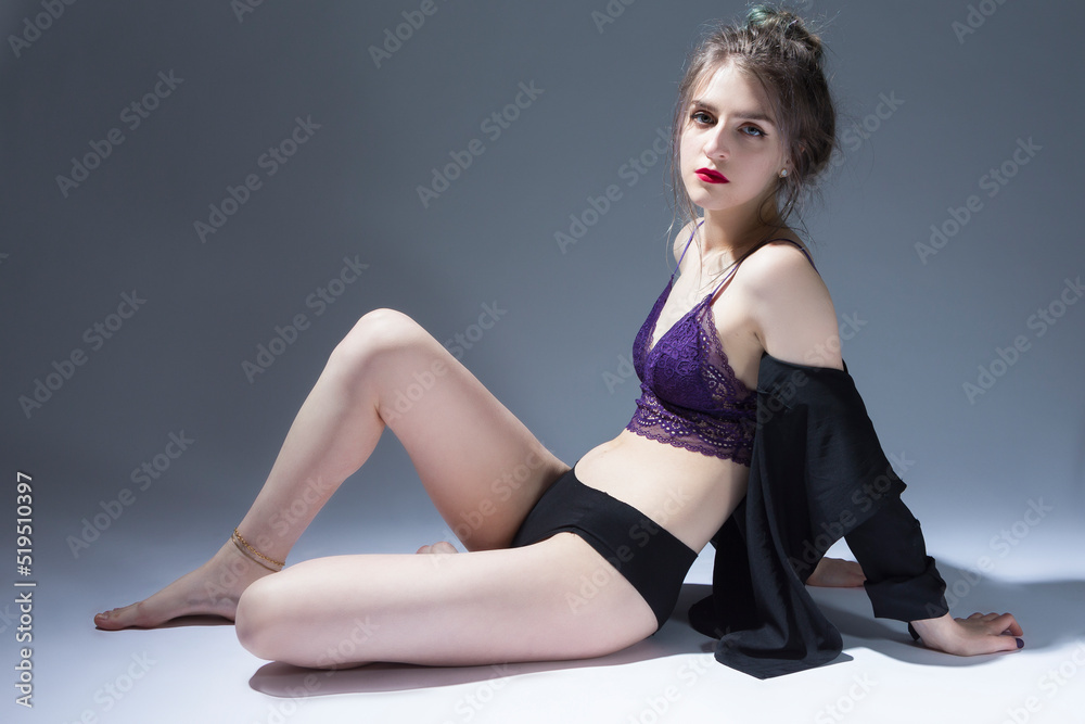Young Caucasian Sensual Girl With Curly Hair Style in Violet Nightie and Black Panties With Shirt Sitting and Posing on Gray Background as Fashion Style Portrait. Young Caucasian Sensual Girl With Curly Hair Style in Violet Nightie and Black Panties With Shirt Sitting and Posing on Gray Background as Fashion Style Portrait.