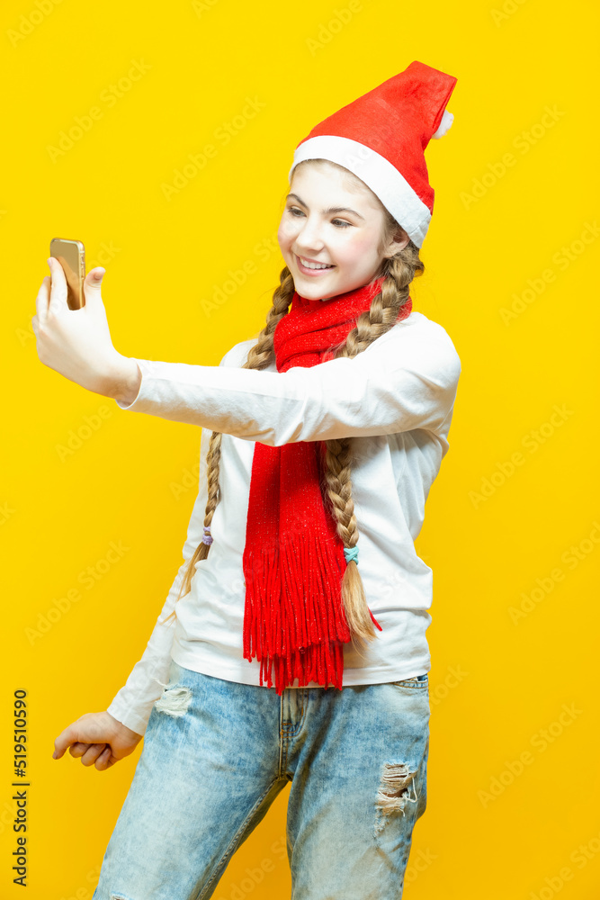 Fototapeta premium Funny Winsome Caucasian Teenager Holding Cellphone Taking Selfie While Smiling and Having Good Time And Making Faces in Christmas Santa Hat Posing Against Yellow Background.