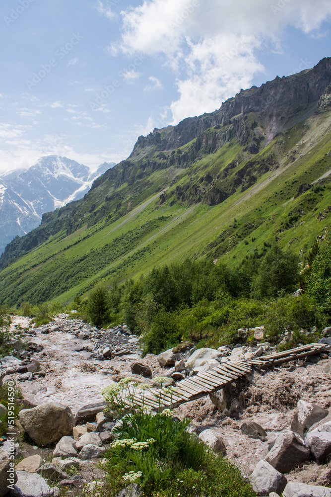 Fototapeta premium A small wooden bridge across the stormy river Terskol and rocky banks among the slopes of mountains and green grass on a bright sunny summer day near Elbrus region