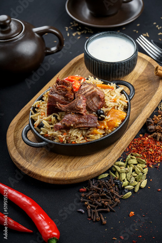 rice pilaf with meat in a round pan on a board with sour cream on a black background with spices, vertical photo 