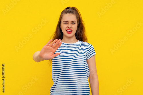 young annoyed woman doing stop sign with serious and confident expression, defense gesture, wears casual white-blue striped t shirt. Indoor studio shot on yellow background