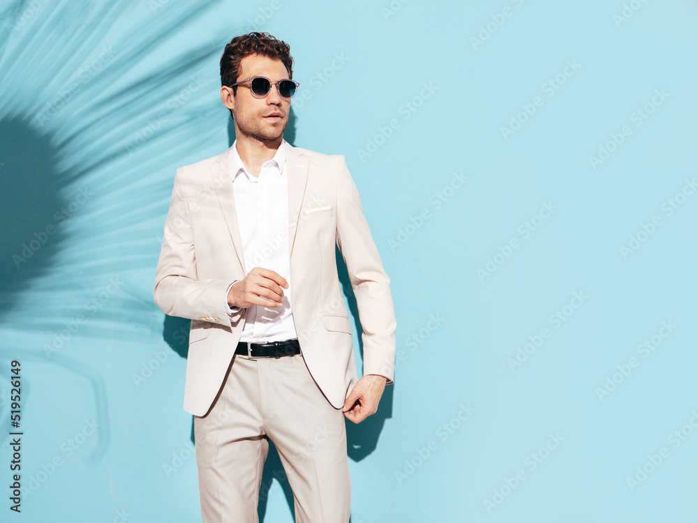 Portrait of handsome confident stylish hipster lambersexual model. Sexy modern man dressed in white elegant suit. Fashion male posing in studio near blue wall in sunglasses Portrait of handsome confident stylish hipster lambersexual model. Sexy modern man dressed in white elegant suit. Fashion male posing in studio near blue wall in sunglasses